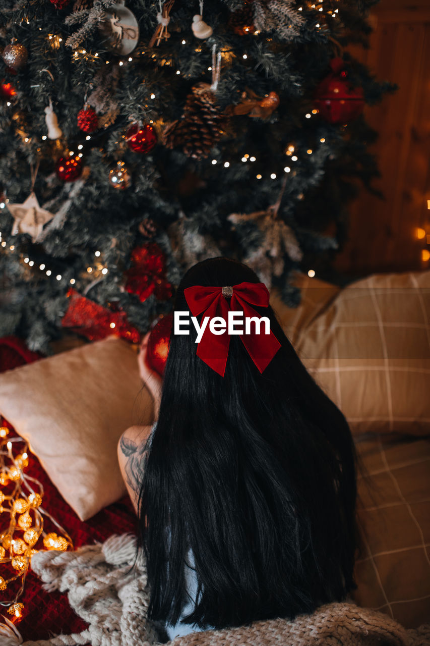 A brunette woman with a red bow in her hair lying near the christmas tree with festive decorations.