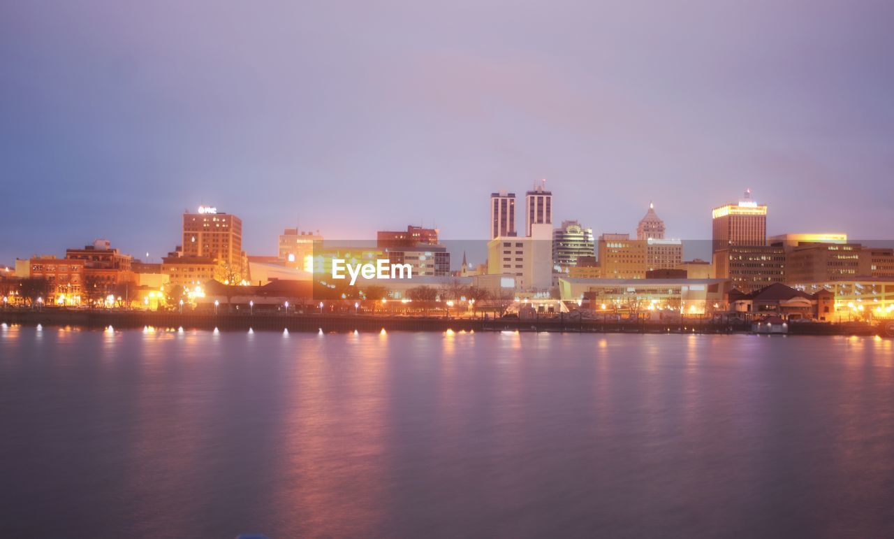 Illuminated buildings by river against sky at night