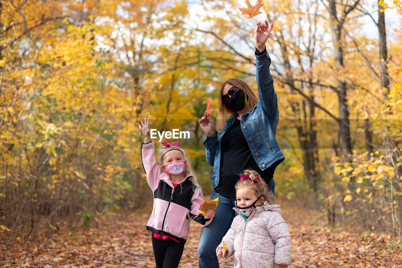 Smiling girls with grandmother wearing mask standing in forest during autumn