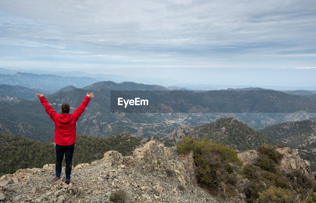 Woman with raised arms standing on rocky top against a cloudy sky enjoying mountain range panorama.
