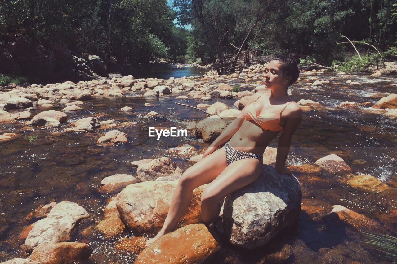 Sensuous woman wearing bikini on rock amidst river at forest