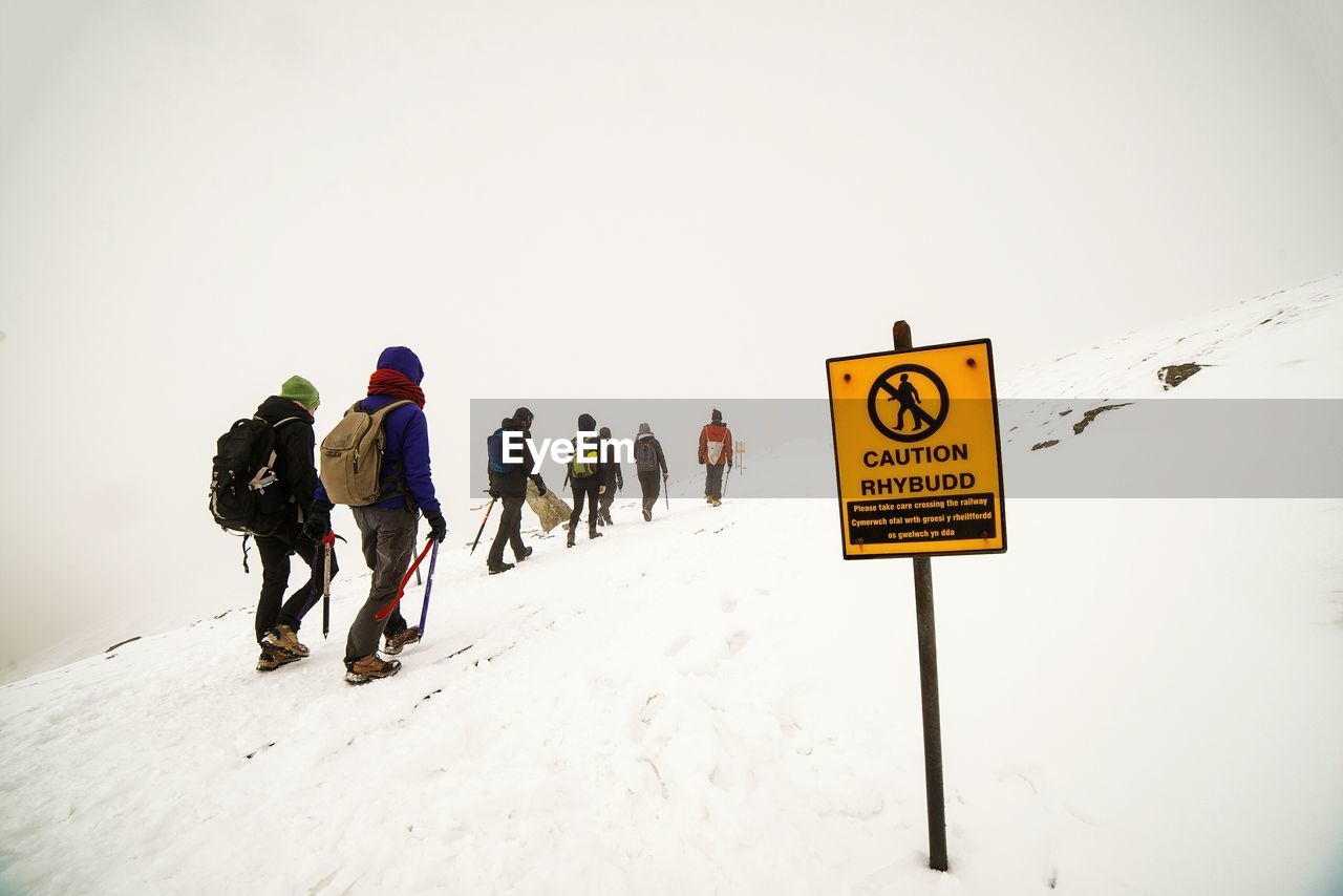 Hikers by sign on snow-covered field against clear sky