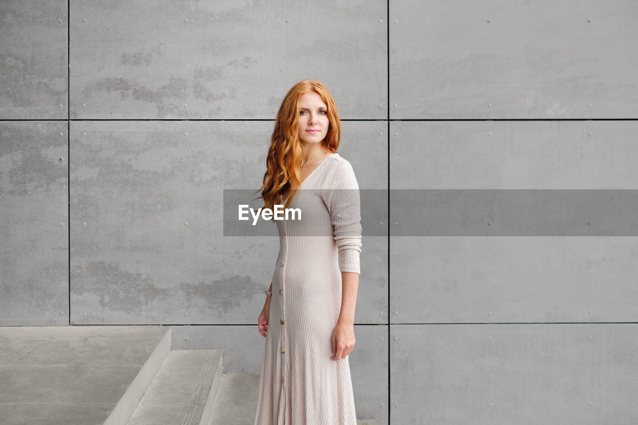 Positive young female with long red hair wearing casual dress and looking at camera while standing against concrete wall