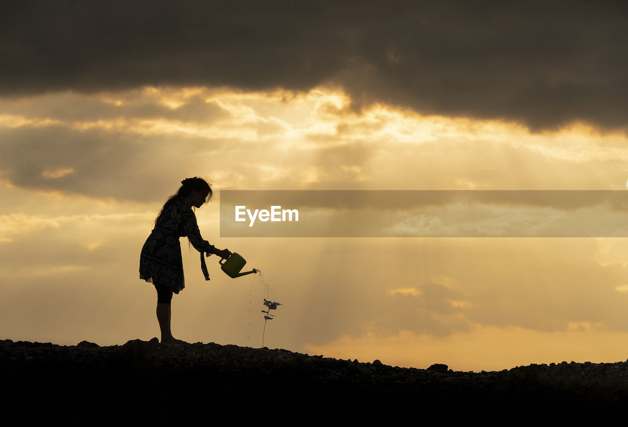 Silhouette woman watering plant on land against sky at sunset