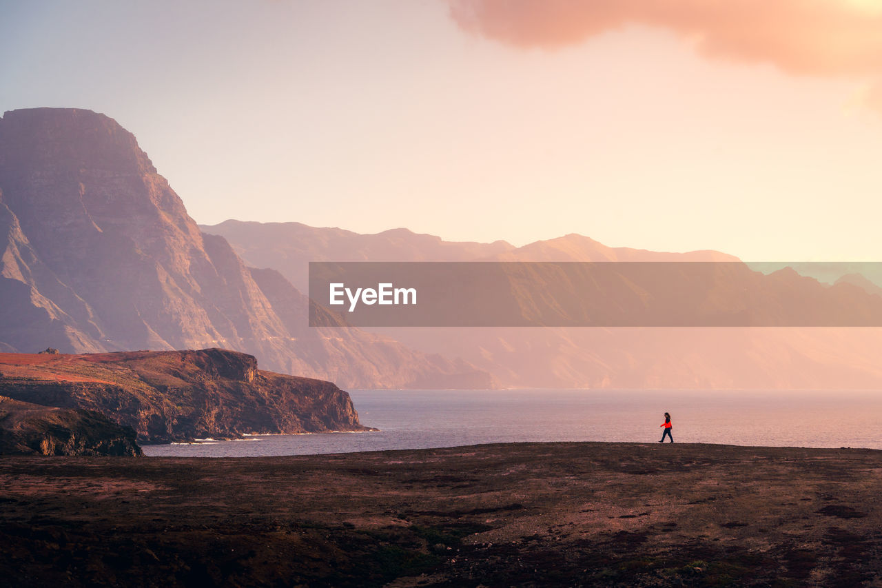 Remote side view of female explorer walking along rocky coast near sea on background of sunset sky and highlands on gran canaria