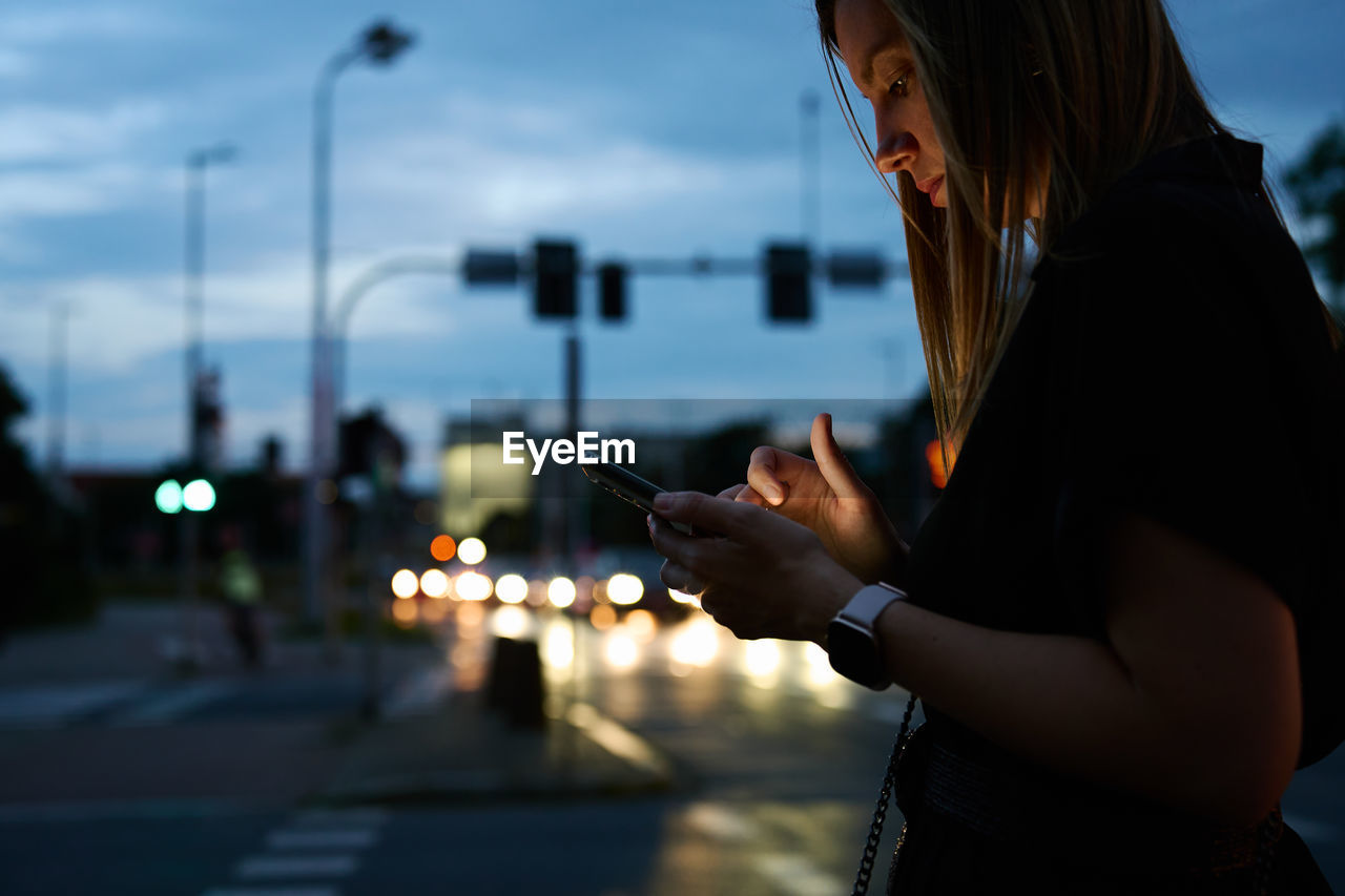 Woman using smartphone on city street with bokeh lights at night. mobile phone in female hands