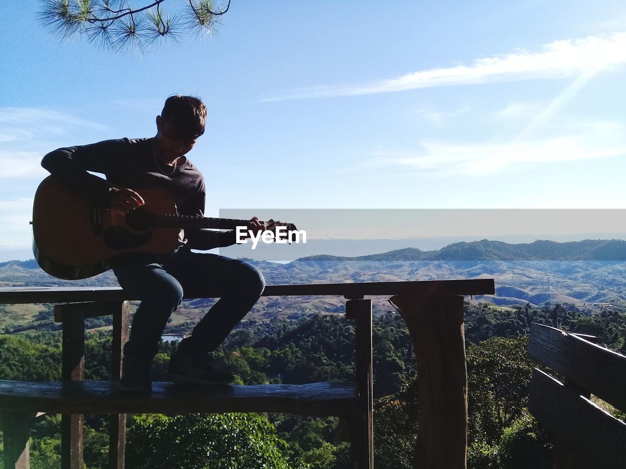 Man playing guitar while sitting on railing against sky