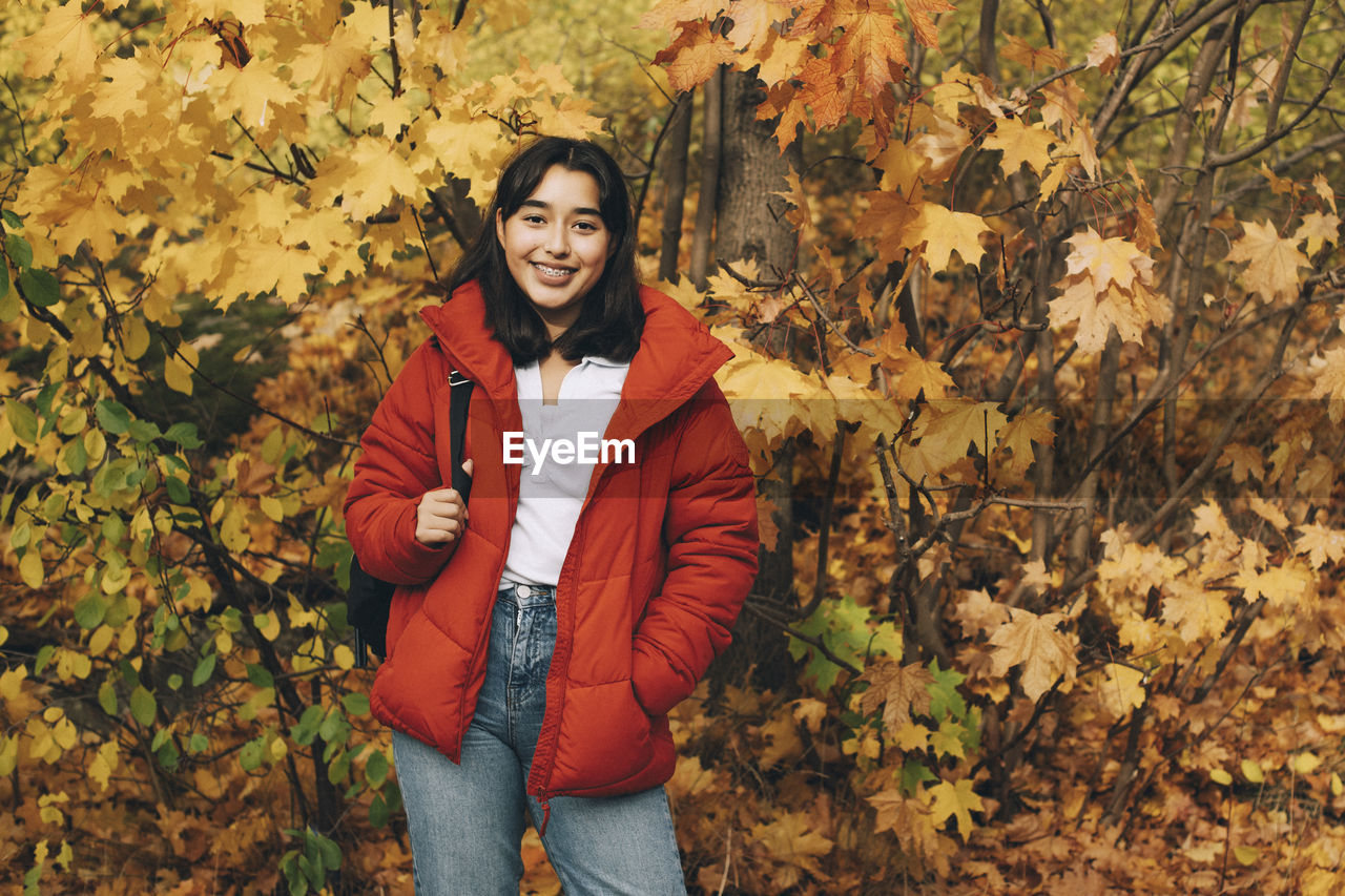 PORTRAIT OF SMILING YOUNG WOMAN STANDING BY LEAVES DURING AUTUMN