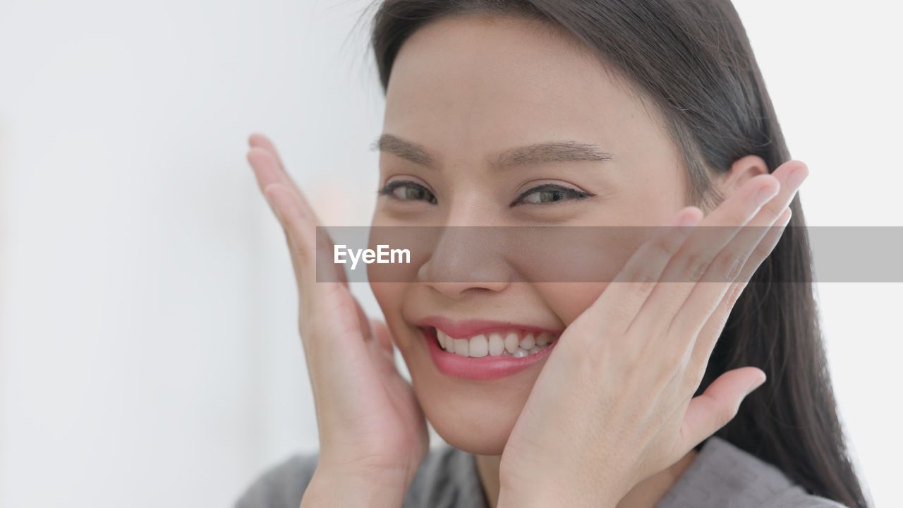 Over a spa background, a young gorgeous woman with a towel on her head cheerfully touches her face.