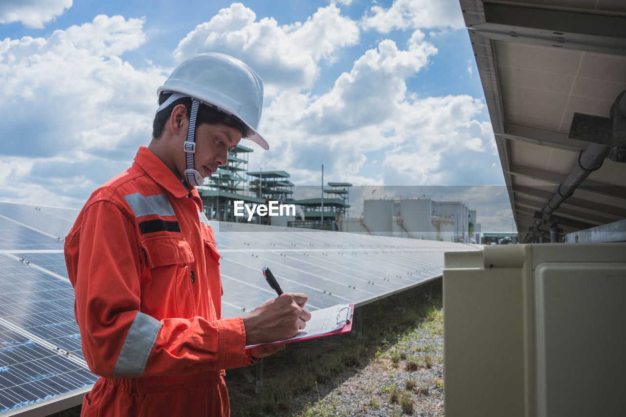 Side view of worker wearing reflective clothing while standing on solar panel against sky