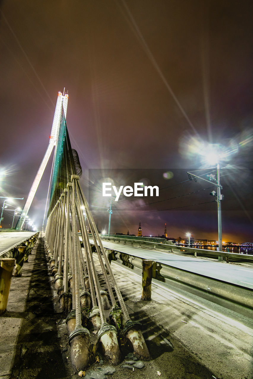 Illuminated street by bridge against sky at night