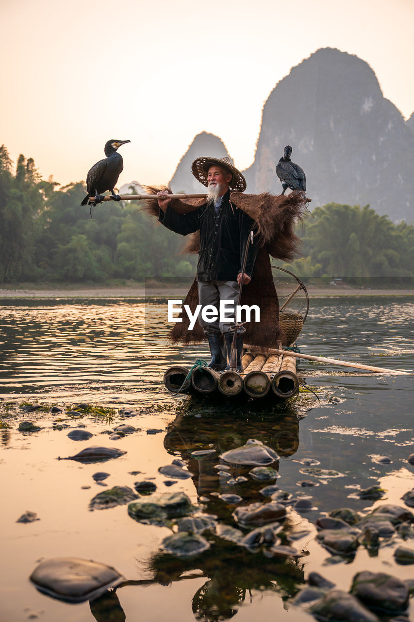 Senior man wearing hat standing on raft against mountain and sky