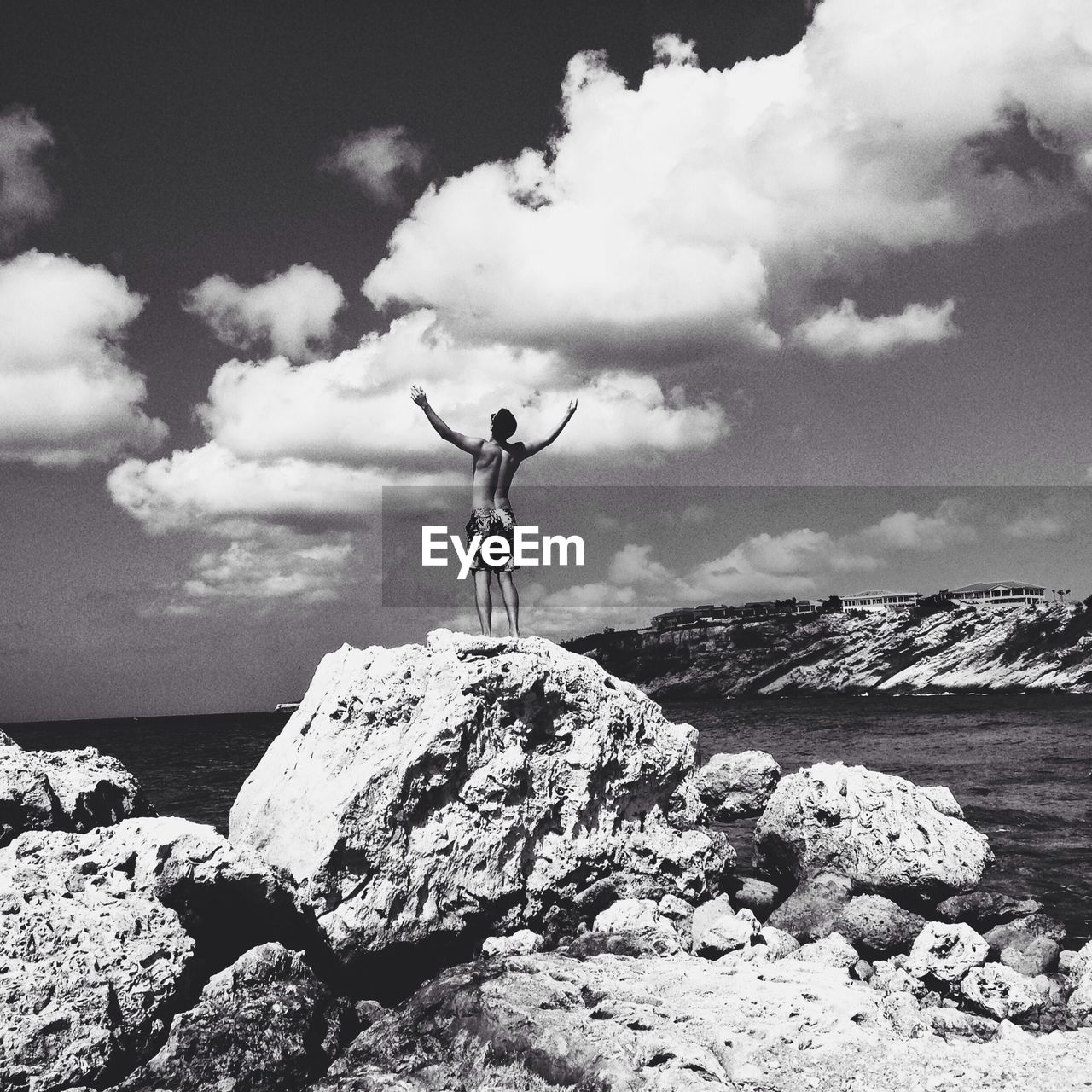 Low angle view of shirtless man with arms outstretched standing on rock by sea against cloudy sky