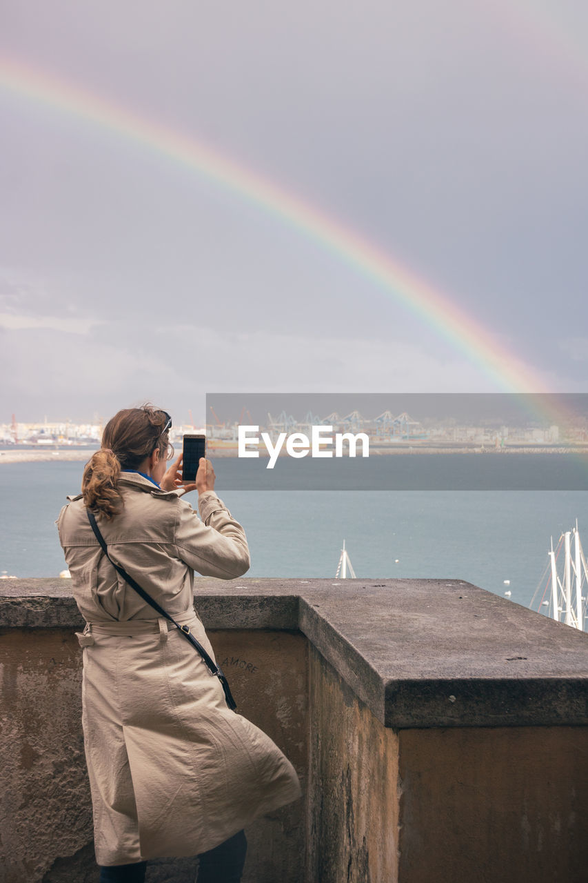 Rear view of woman photographing rainbow over sea against sky