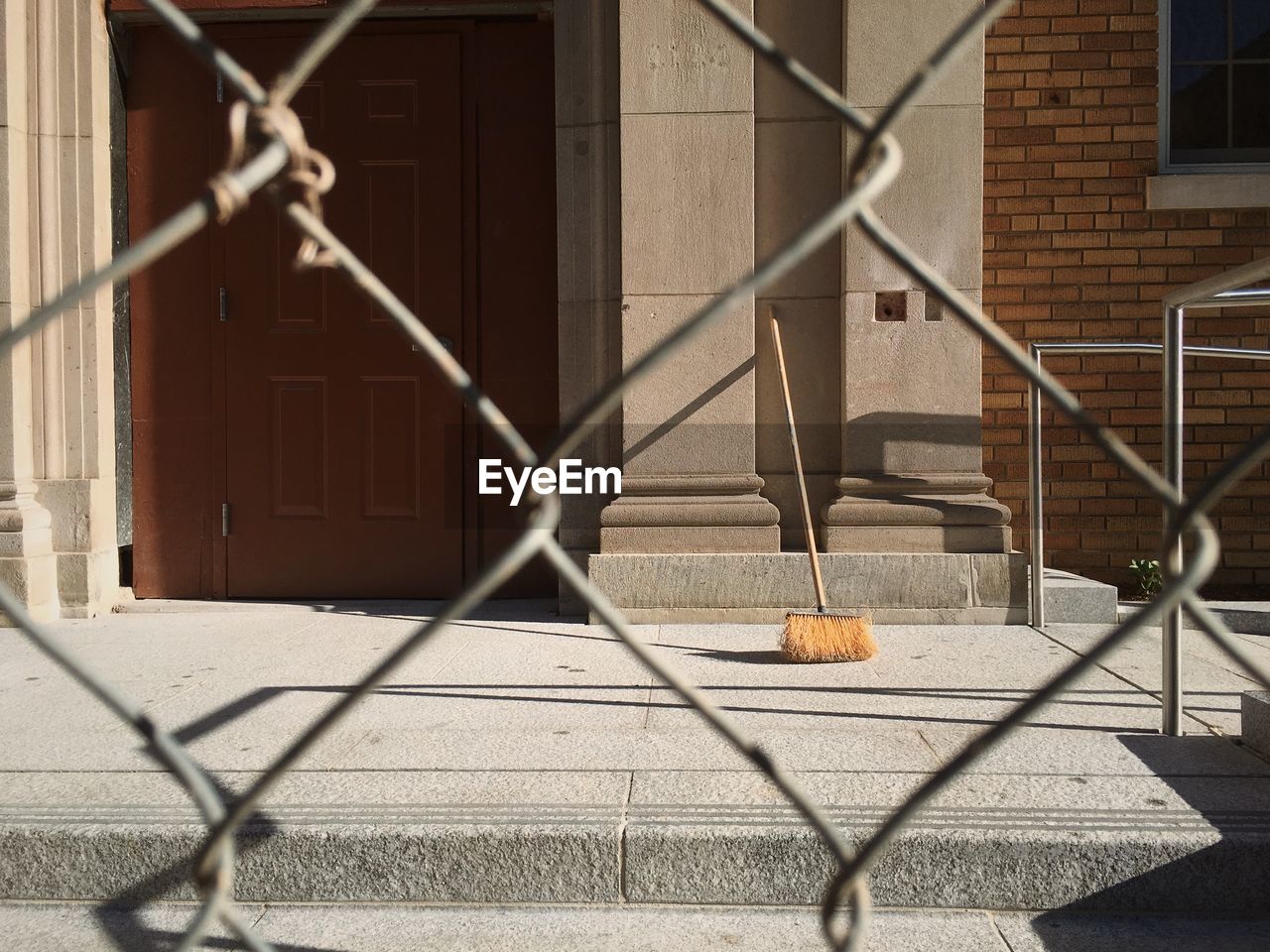 Broom on sidewalk against building seen through chainlink fence