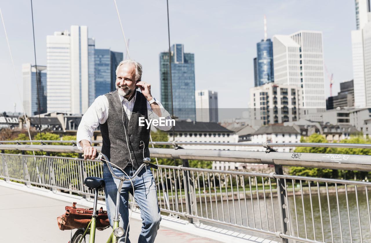 Smiling mature man with earbuds and bicycle crossing bridge in the city