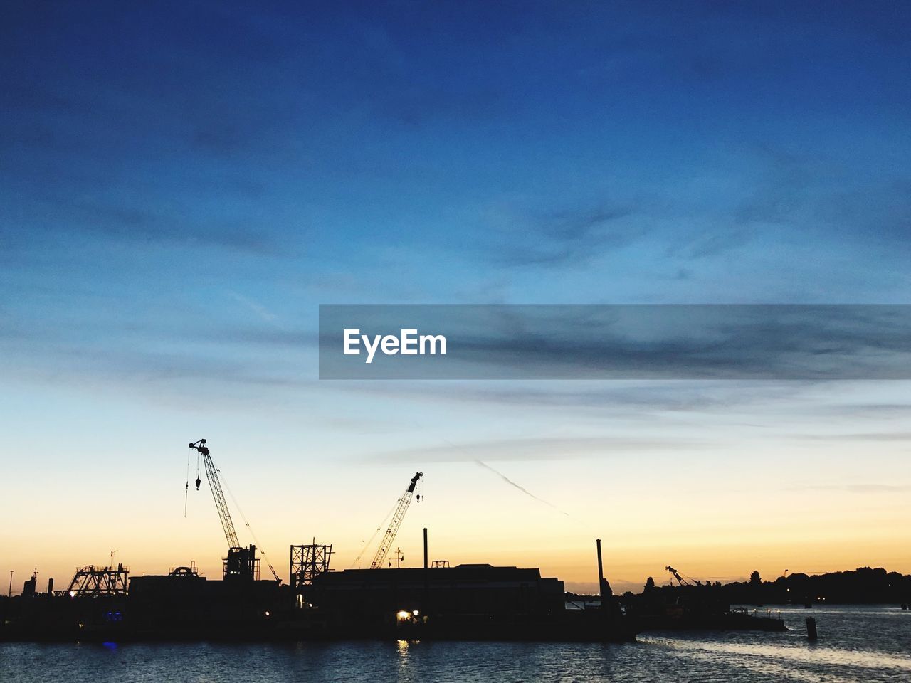 Silhouette cranes at harbor against blue sky during sunset