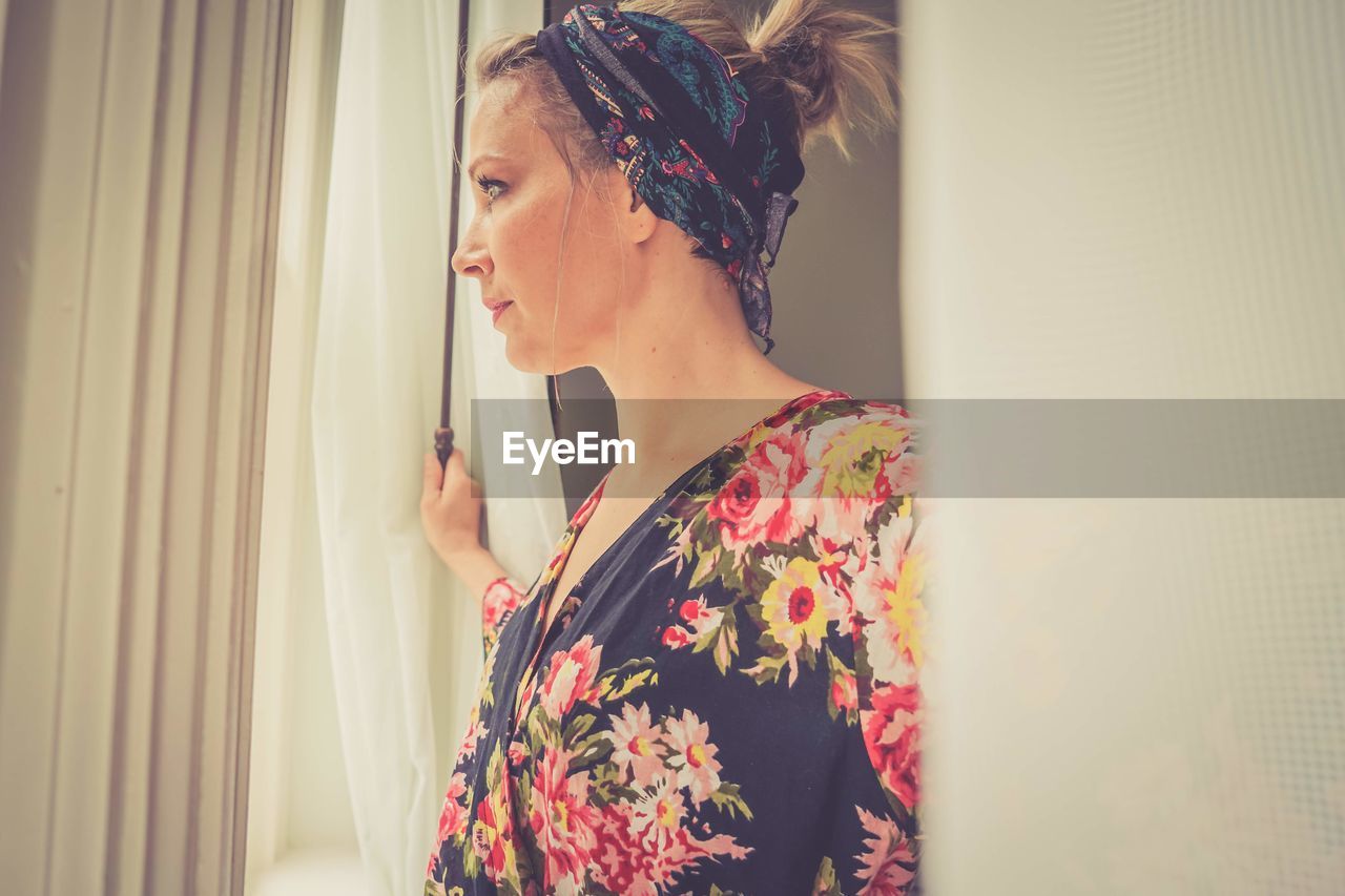 Young woman looking away while standing by window at home