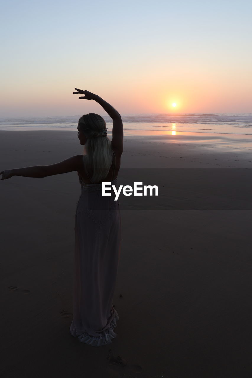 REAR VIEW OF WOMAN ON BEACH DURING SUNSET