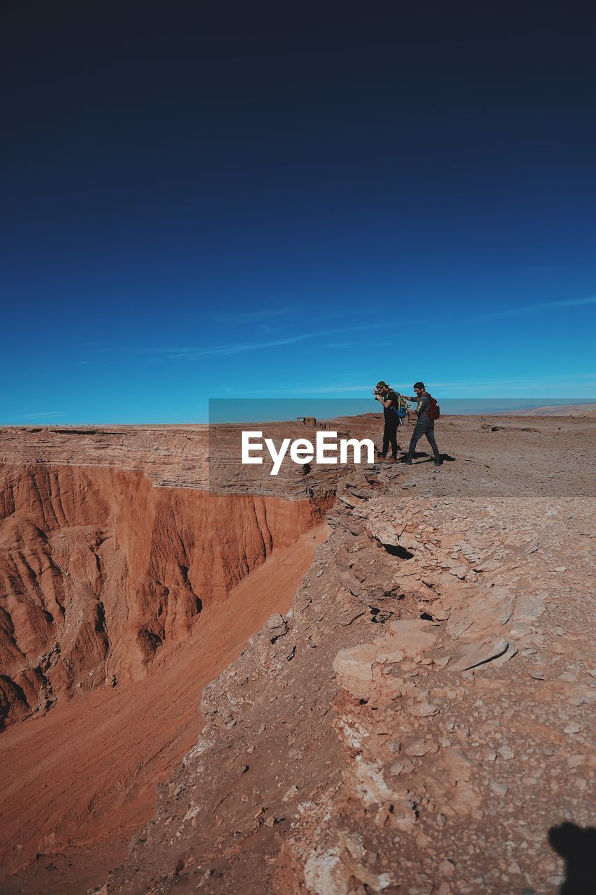 Man photographing through camera with friend while standing on cliff against blue sky