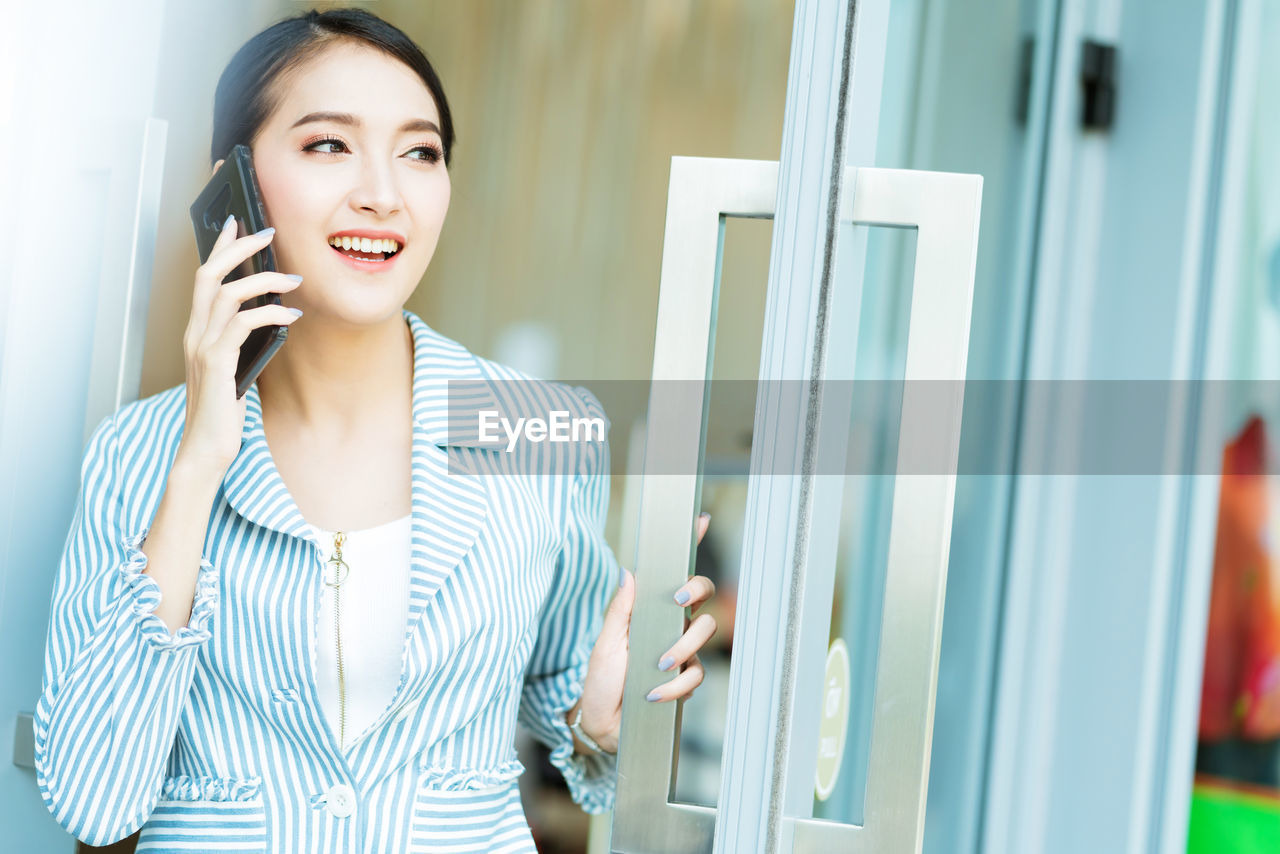 SMILING YOUNG WOMAN LOOKING THROUGH SMART PHONE WHILE STANDING ON LAPTOP IN MIRROR