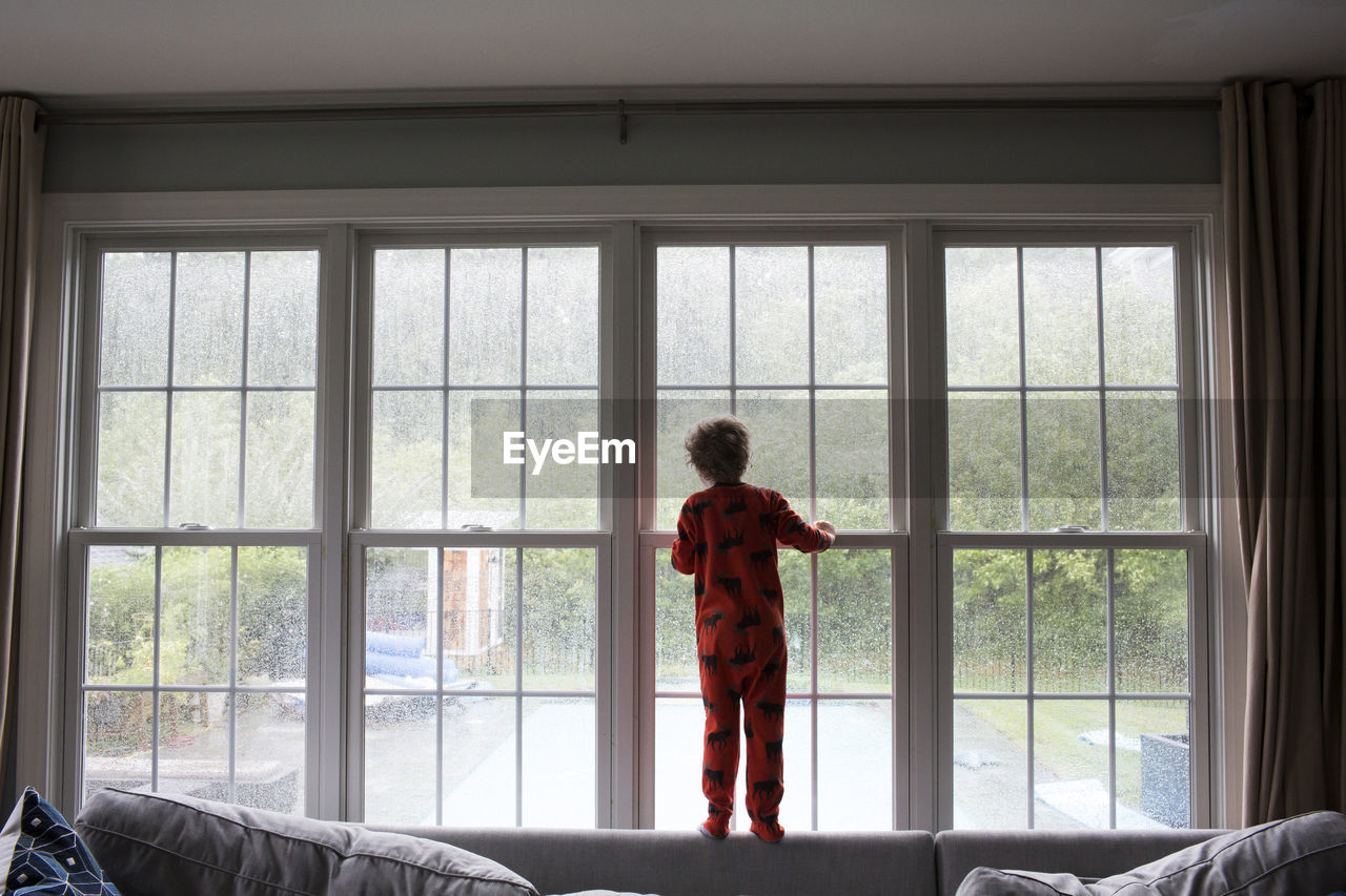 Small boy looks out large living room window at rainy back yard