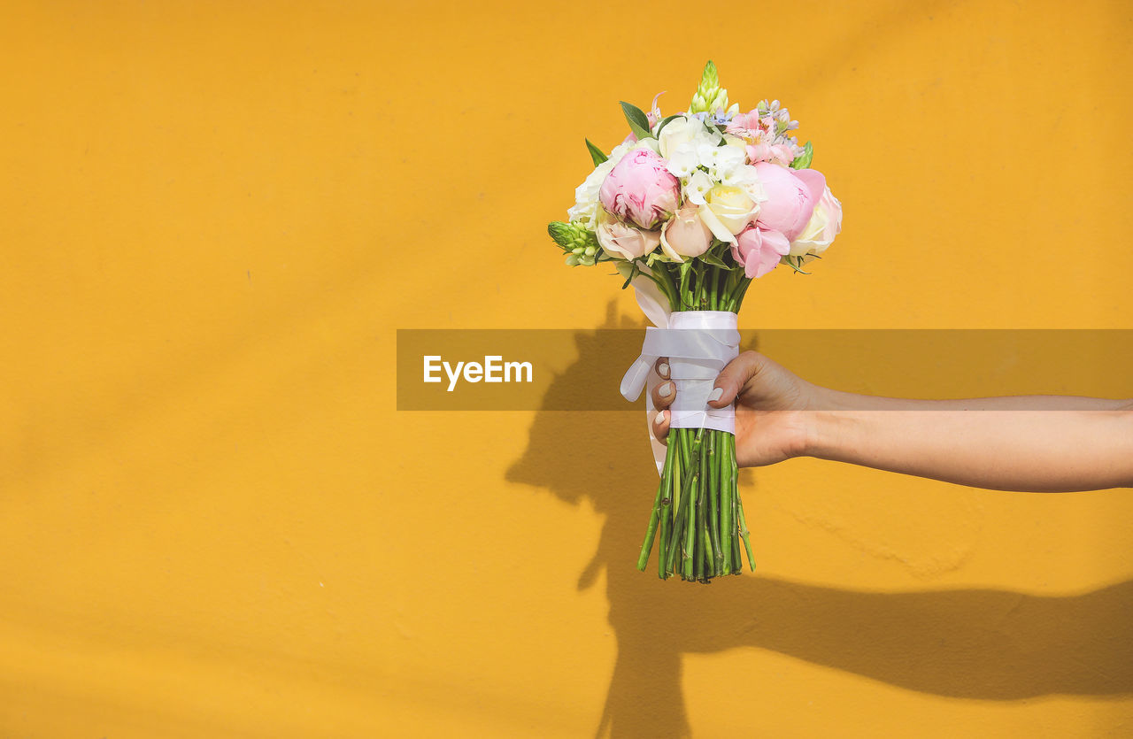 Cropped hand of woman holding bouquet against yellow wall