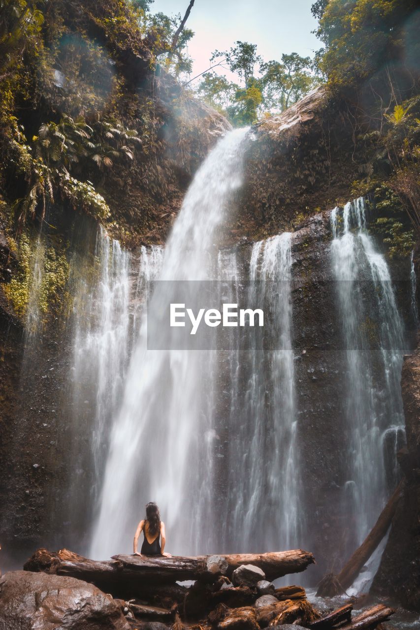 Rear view of woman sitting against waterfall in forest