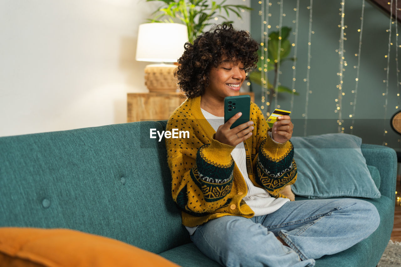 portrait of smiling young woman using mobile phone while sitting on sofa at home