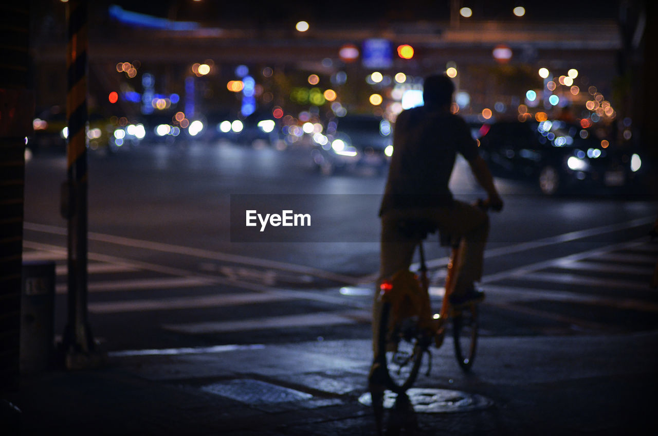 Rear view of man riding bicycle on city street at night