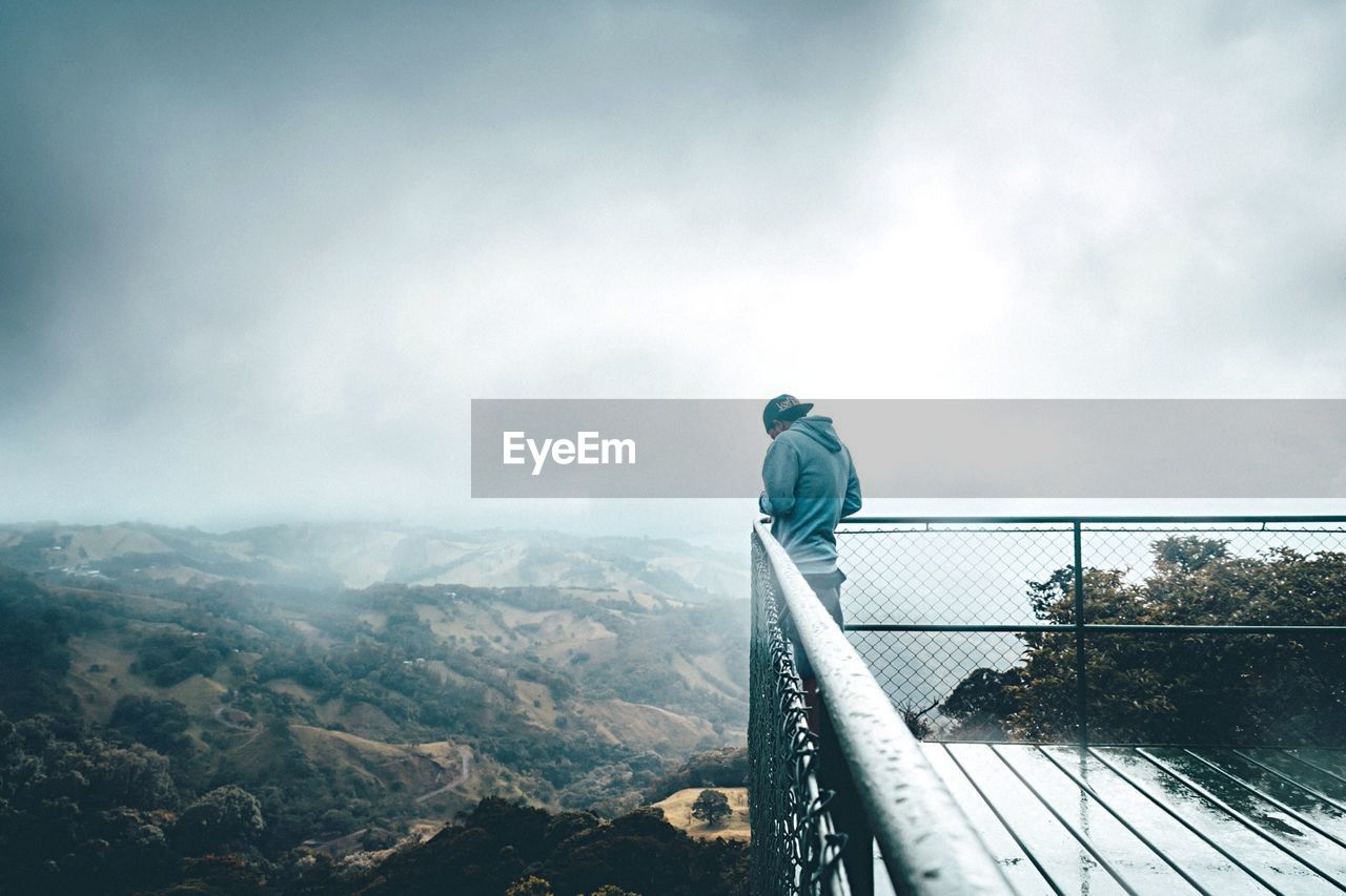 Man standing at observation point by landscape against cloudy sky