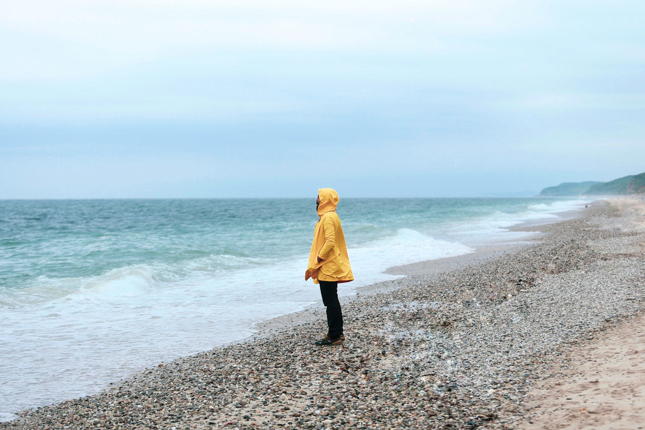 Side view of man wearing raincoat while standing at beach against sky