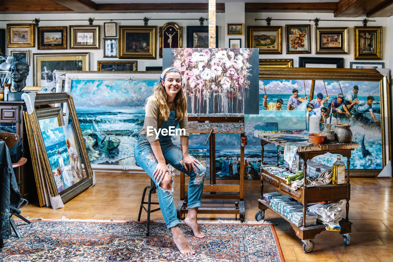 Cheerful female artist in denim overall looking at camera while sitting near easel and painting in creative atelier