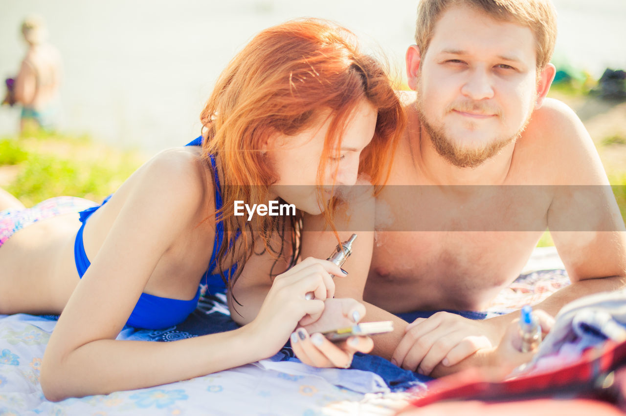 Portrait of smiling man with girlfriend holding electronic cigarette lying at beach