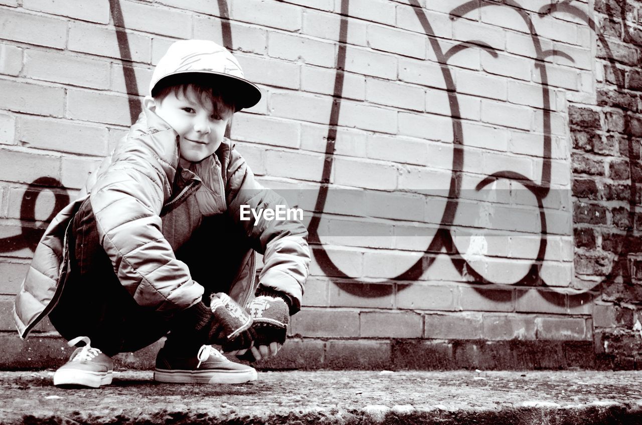 Low angle portrait of smiling boy crouching on sidewalk against wall