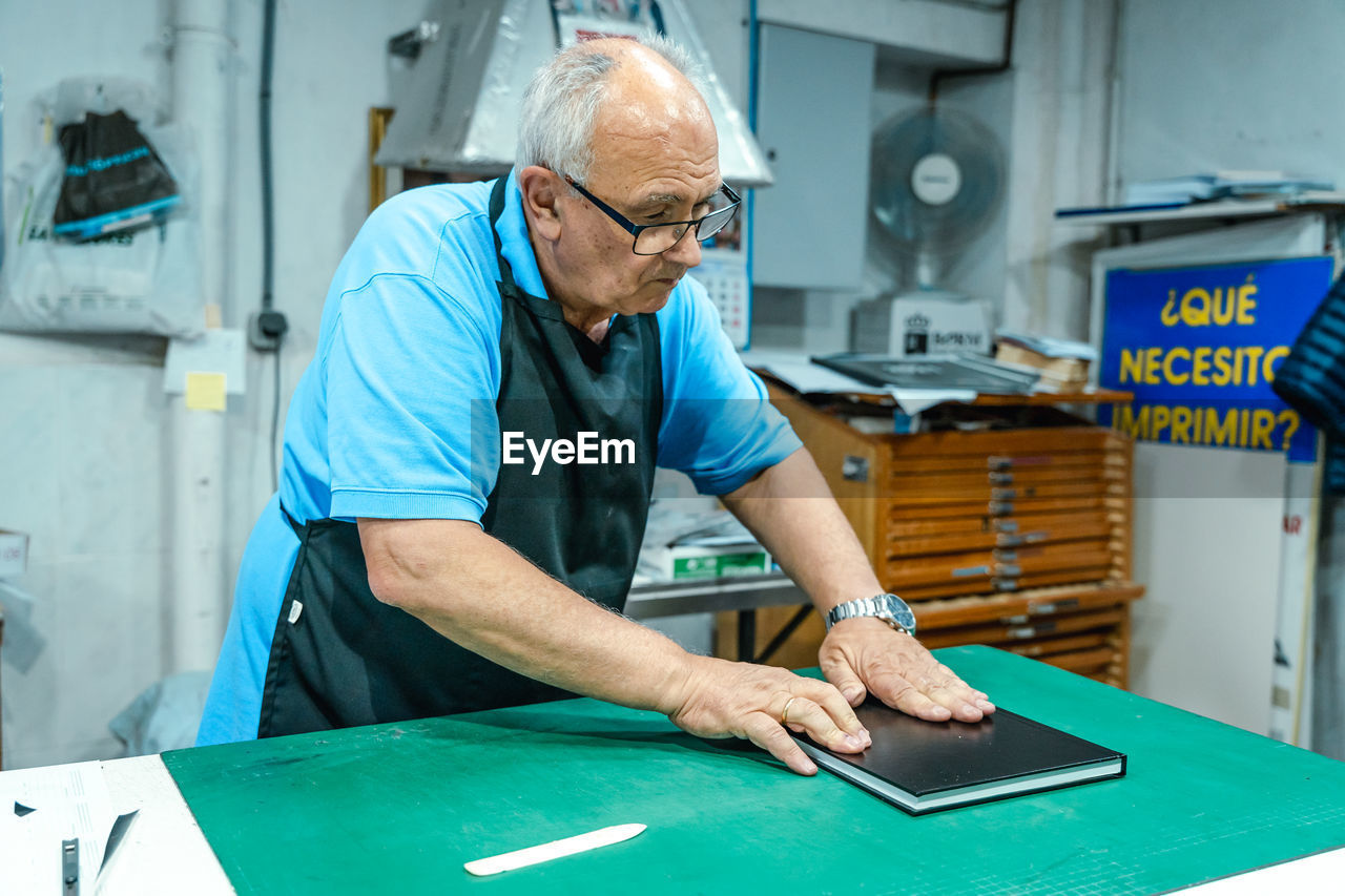 Middle aged male artisan in apron making binding for book standing at table in printing workshop