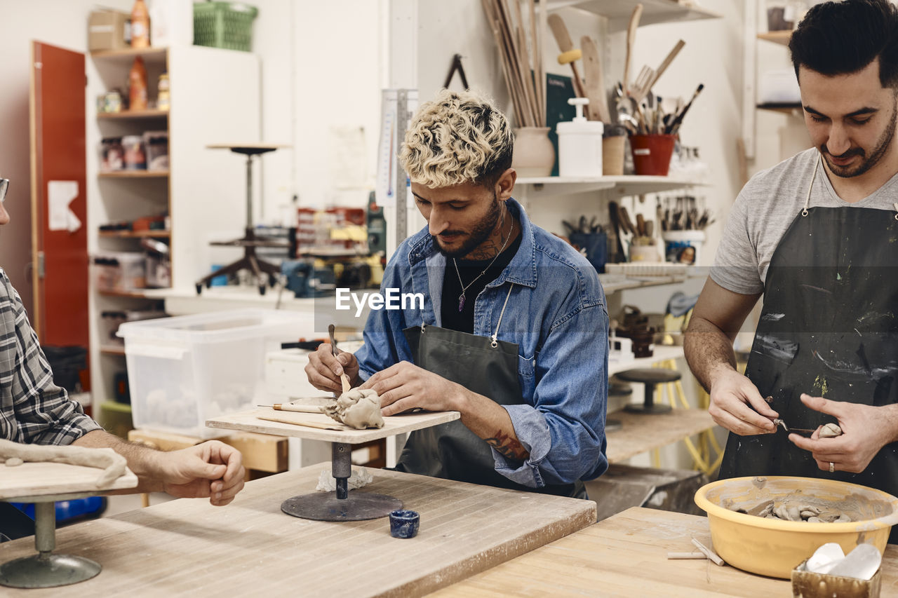 Young man using work tool by friend molding clay while standing at table in art class