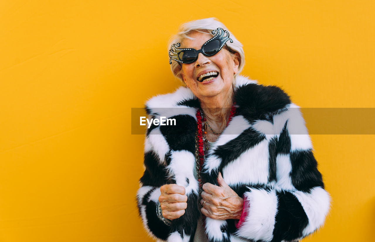 Portrait of senior woman laughing while standing against yellow background