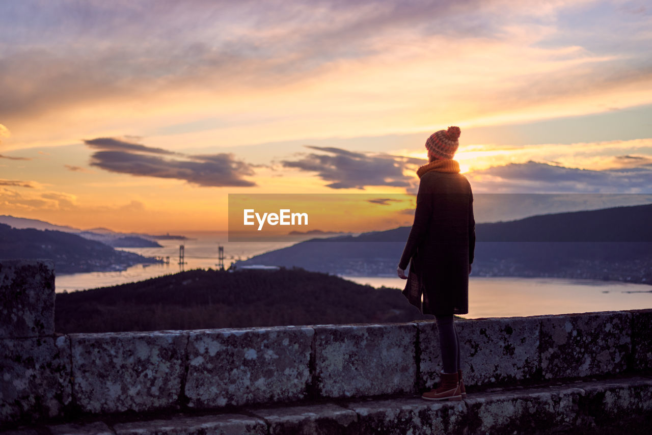 Rear view of woman standing on retaining wall during sunset