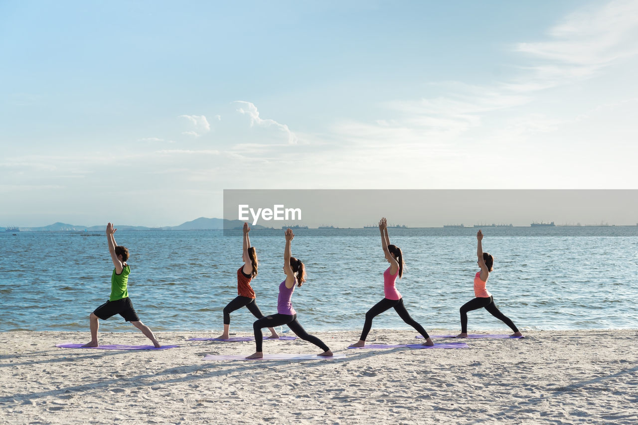 Young friends practicing yoga in warrior position on shore at beach during sunny day