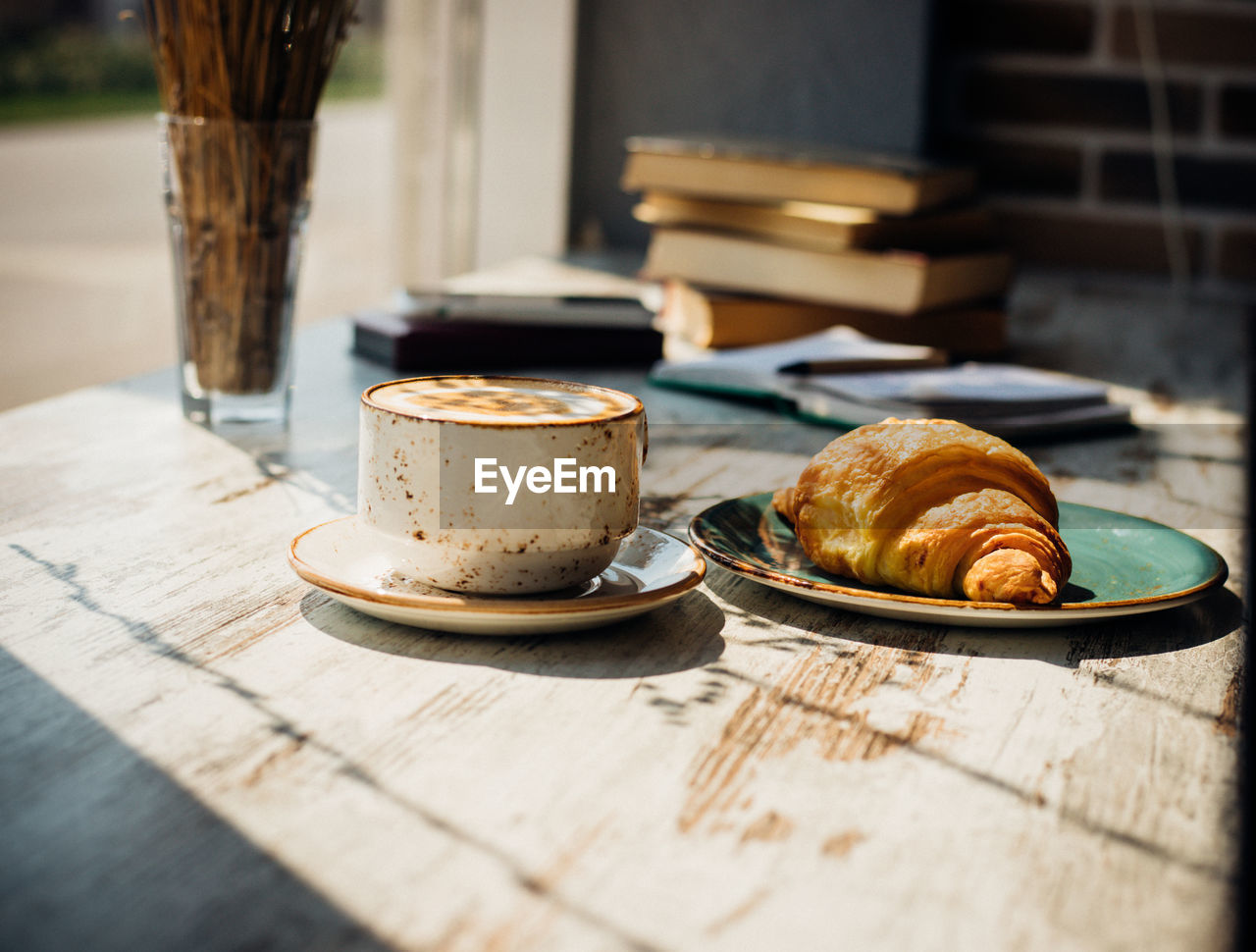 Cappuccino and croissant are lying on the table in a cafe against the background of a window.