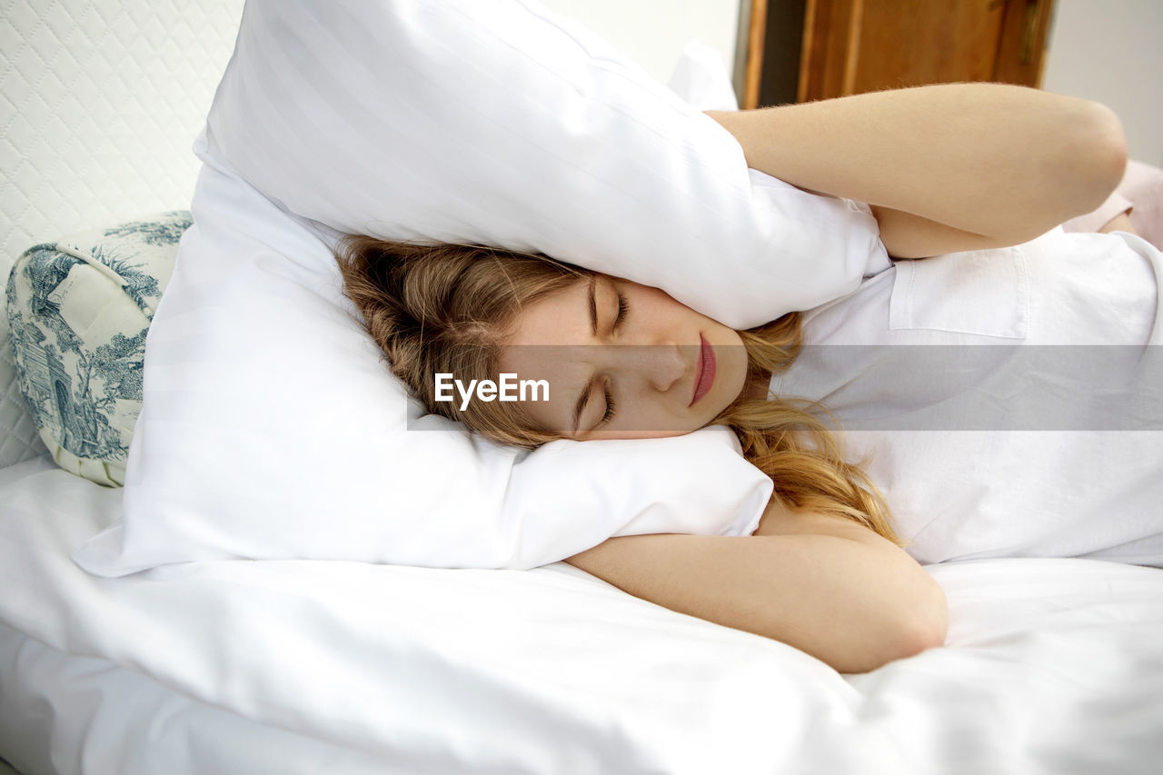 Woman covering ears with pillow while sleeping on bed