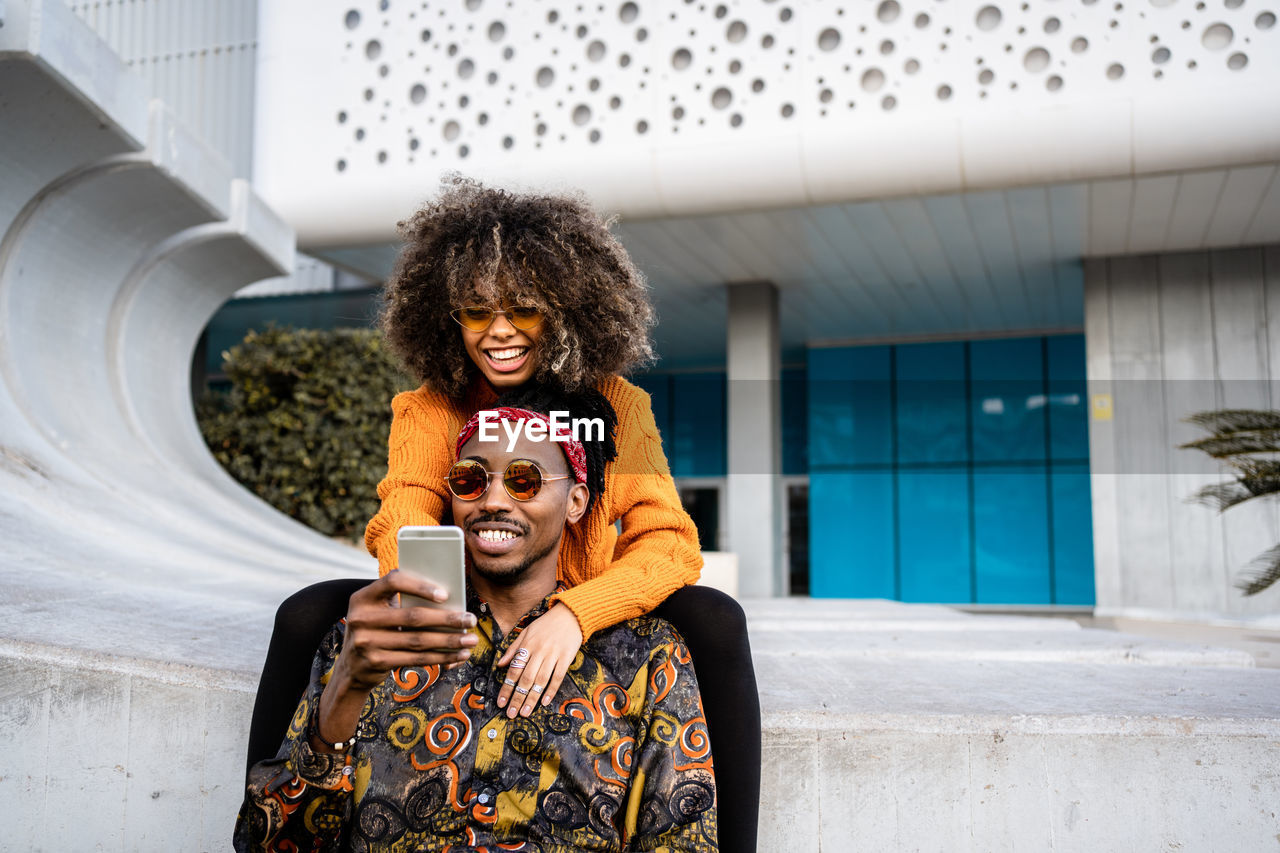 Modern black man using mobile phone with charming ethnic woman with afro hairstyle embracing while chilling on modern bench in city