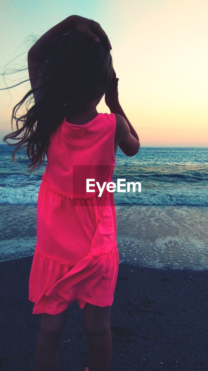 REAR VIEW OF WOMAN STANDING AT BEACH DURING SUNSET