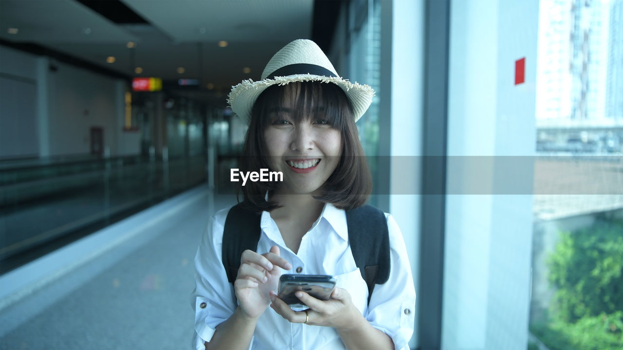 PORTRAIT OF SMILING YOUNG WOMAN USING MOBILE PHONE IN LAPTOP