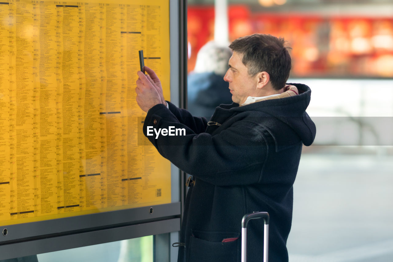 Side view of mature man photographing at arrival departure board at station