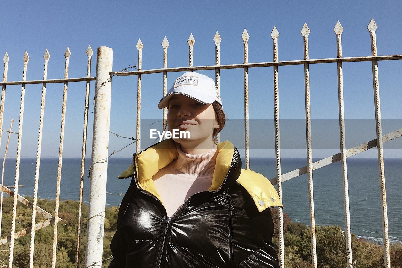fence, one person, protection, security, sky, nature, clothing, leisure activity, sea, adult, water, young adult, standing, lifestyles, railing, day, waist up, women, front view, human face, casual clothing, person, portrait, fashion, outdoors, looking, jacket, hat, fashion accessory, sunlight, architecture, glasses, clear sky, metal, looking away, three quarter length, smiling