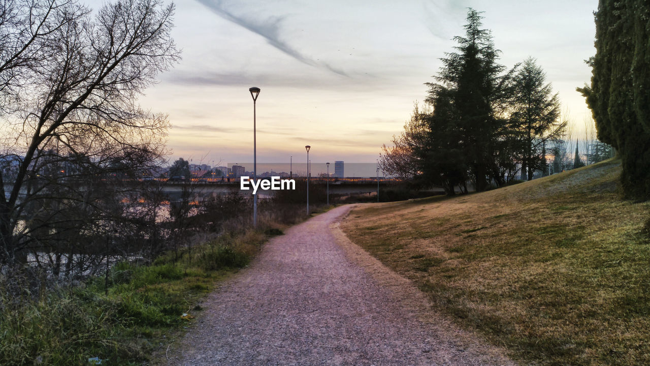 WALKWAY AMIDST TREES AGAINST SKY AT SUNSET