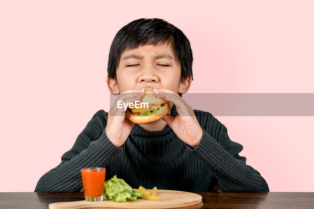 Cute asian boy eating a delicious hamburger with happiness on pink background.