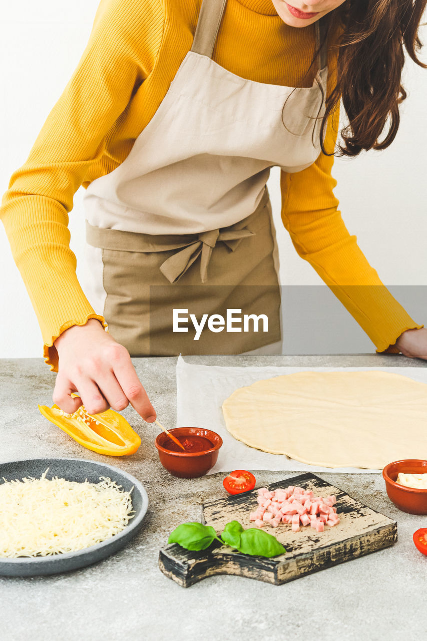 Caucasian girl in an apron at the table with ingredients and dough mixes the sauce for making pizza 